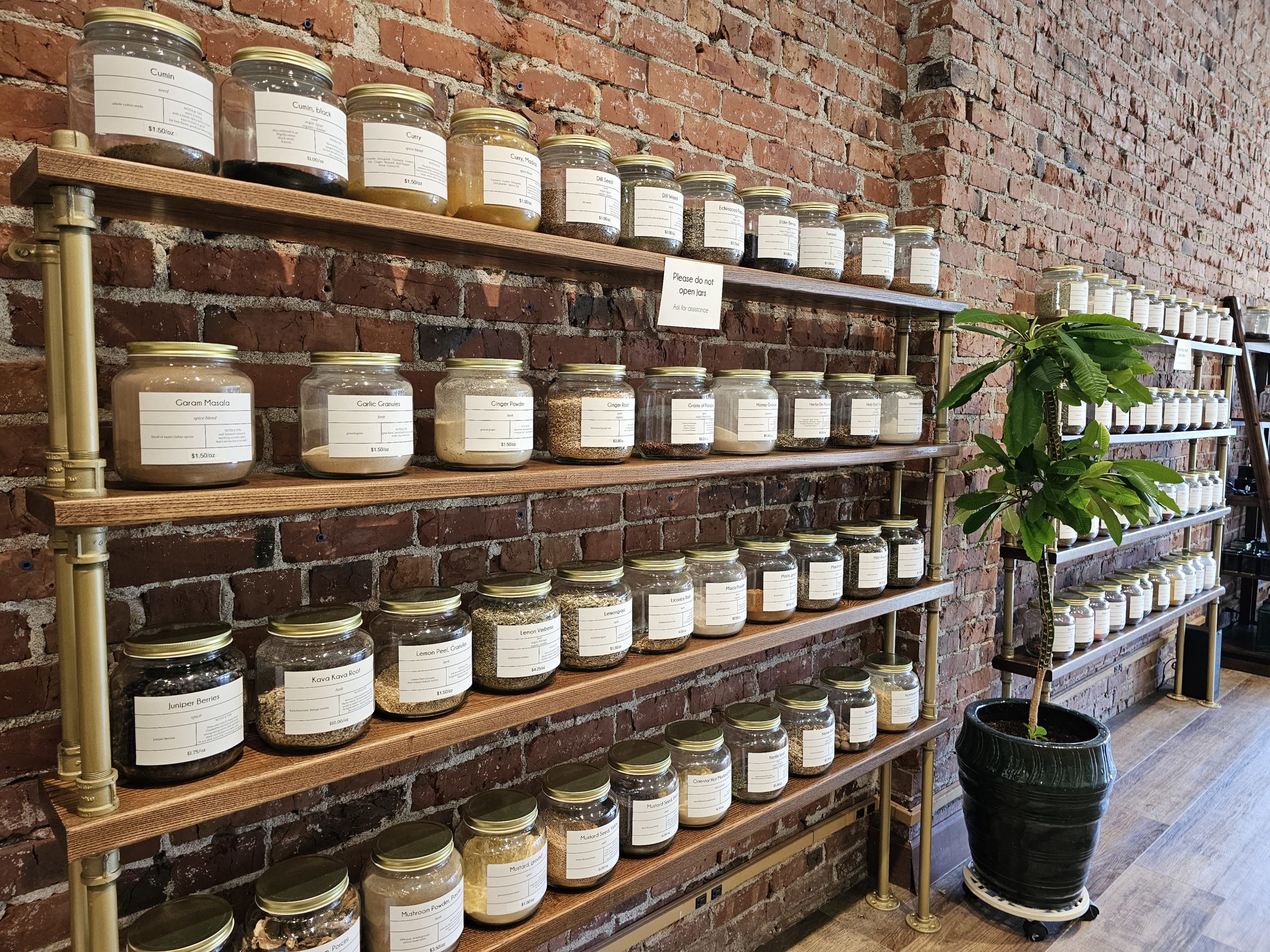 a shelf made of wood and pipe, full of glass jars with spices, in front of a brick wall