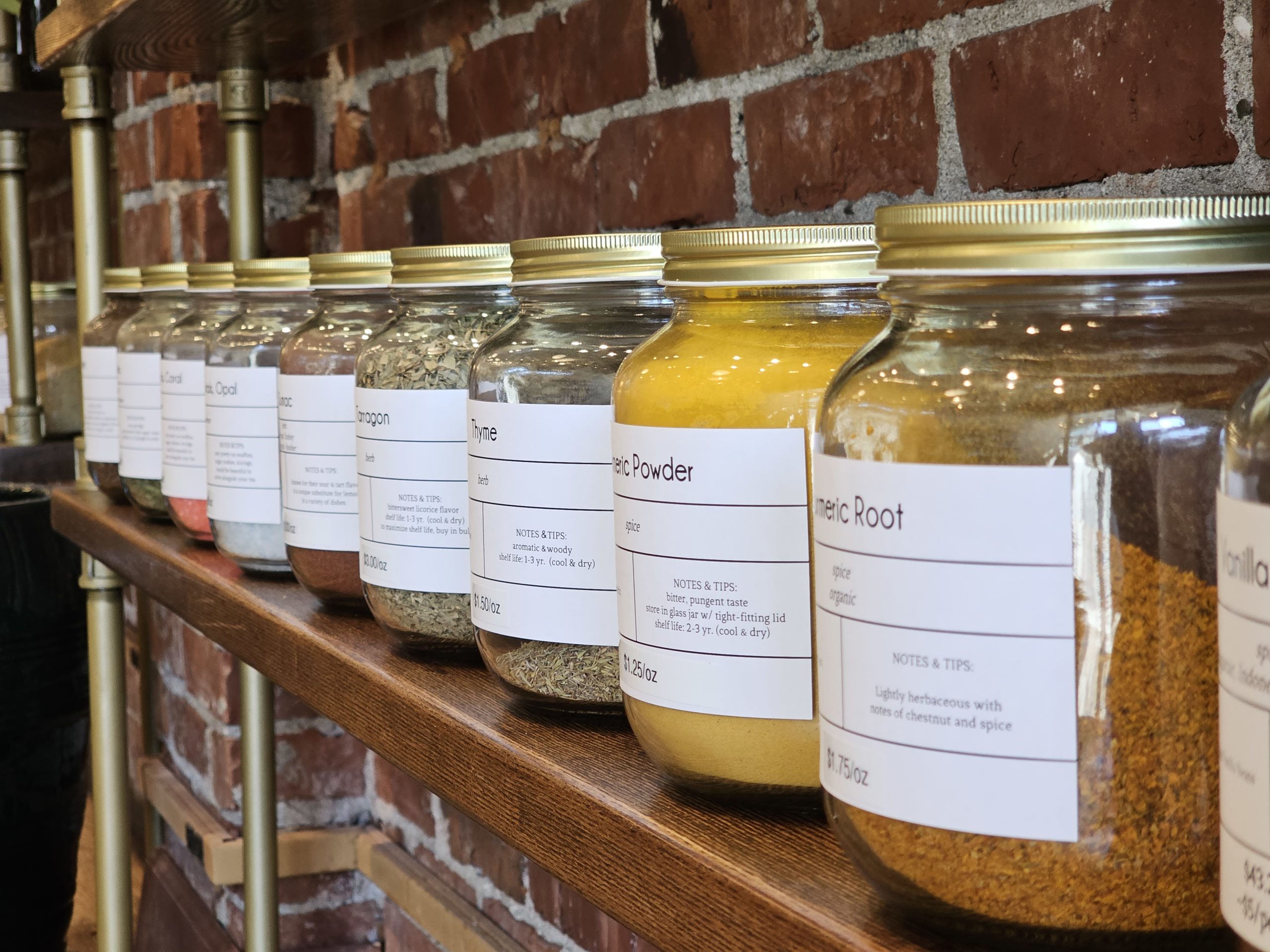 a row of glass jars full of colorful spices, with white labels and gold lids, sitting on a wooden shelf in front of a brick wall