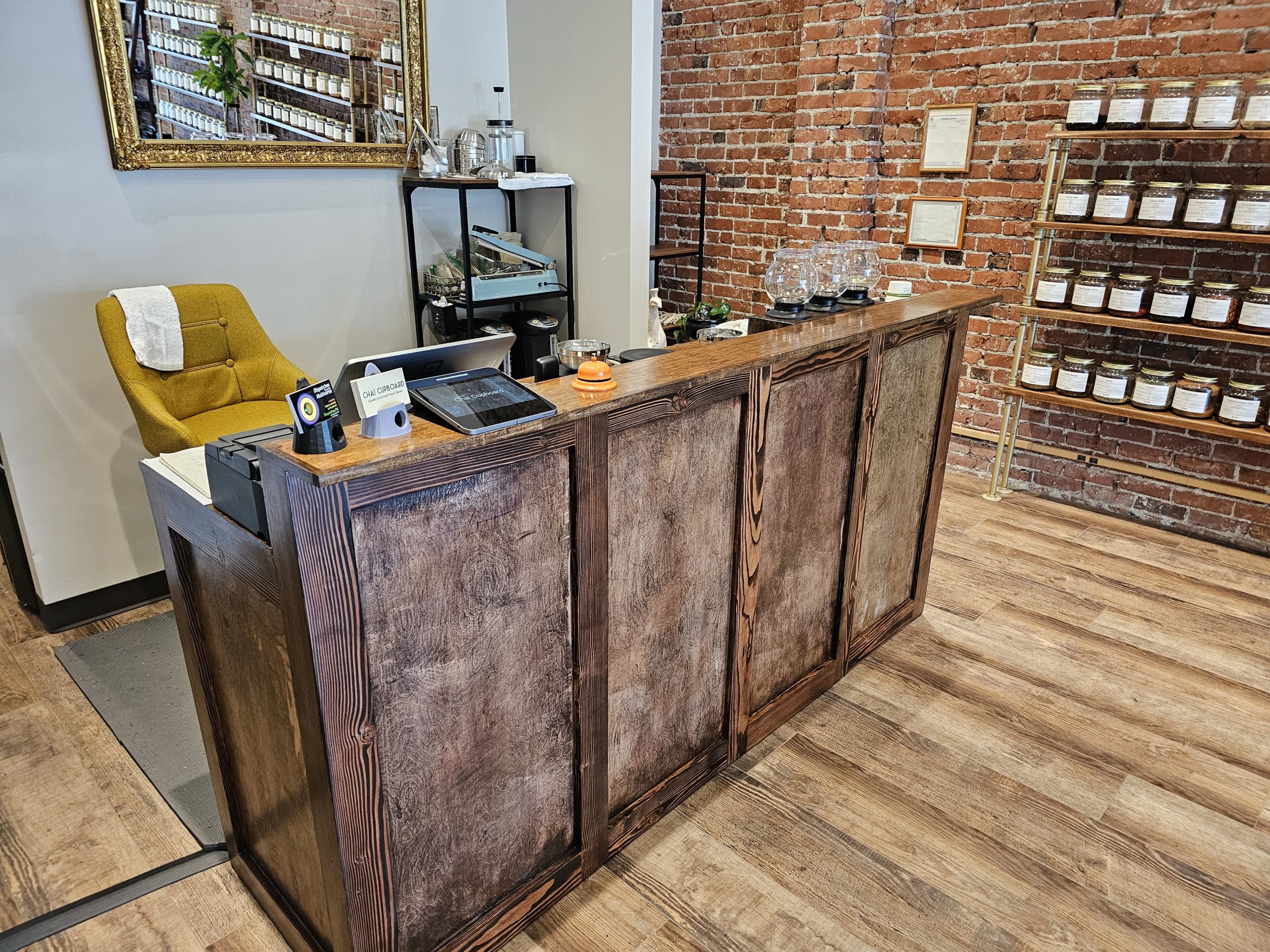 a custom-built wooden counter holding a register and some tea brewers, in a shop with brick walls and a shelf with jars in the background